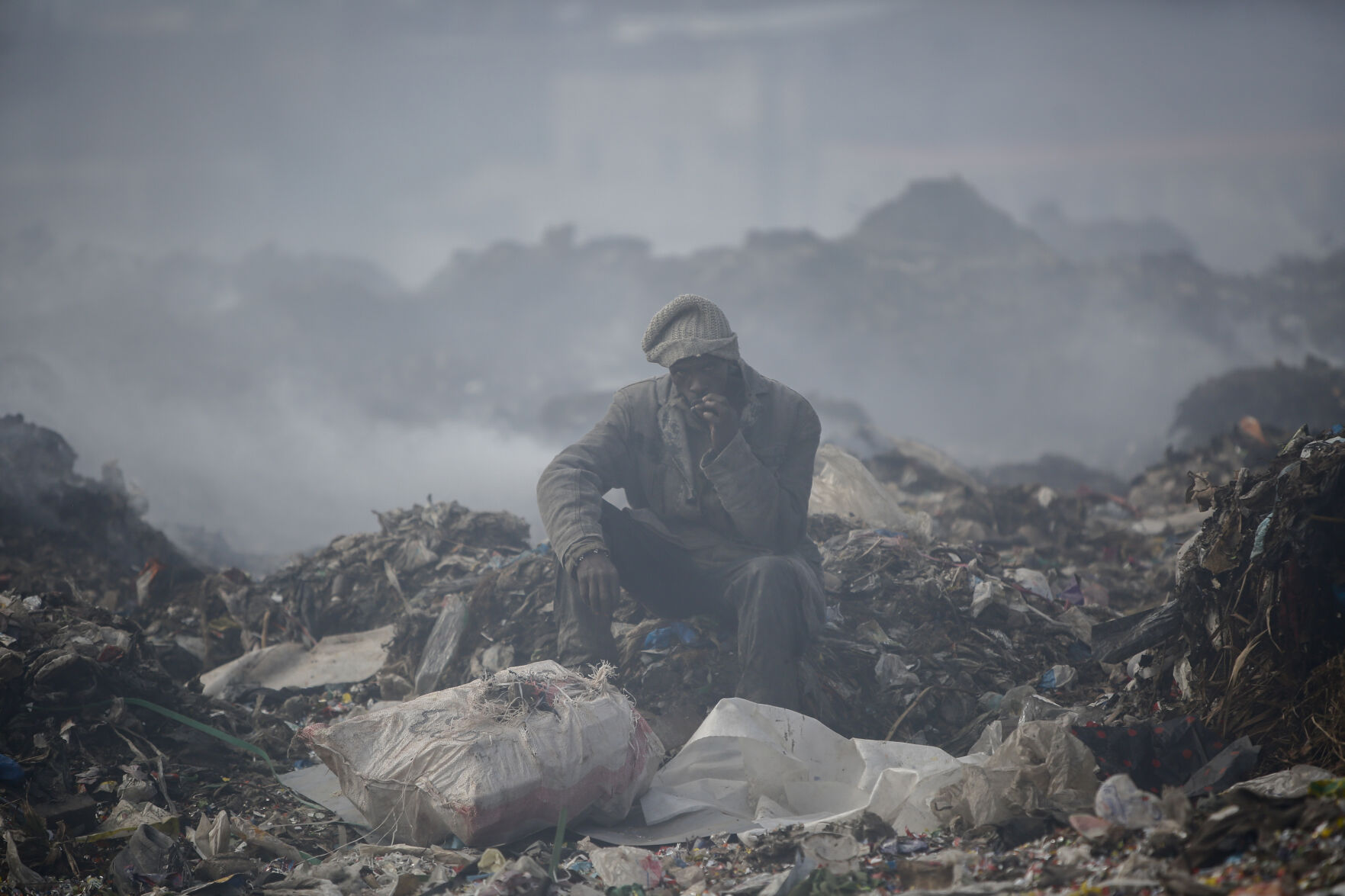 <p>FILE - A man who scavenges recyclable materials for a living rests to smoke a cigarette on a mountain of garage amidst smoke from burning trash at Dandora, the largest garbage dump in the capital Nairobi, Kenya, Tuesday, Sept. 7, 2021.</p>